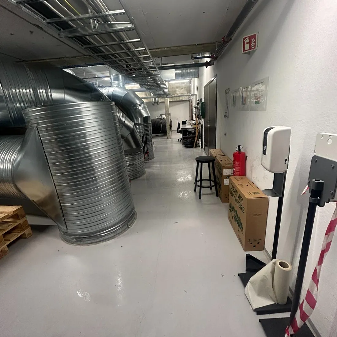 Teknisk rom med store ventilasjonsrør og ledninger, paller og utstyr langs veggene.