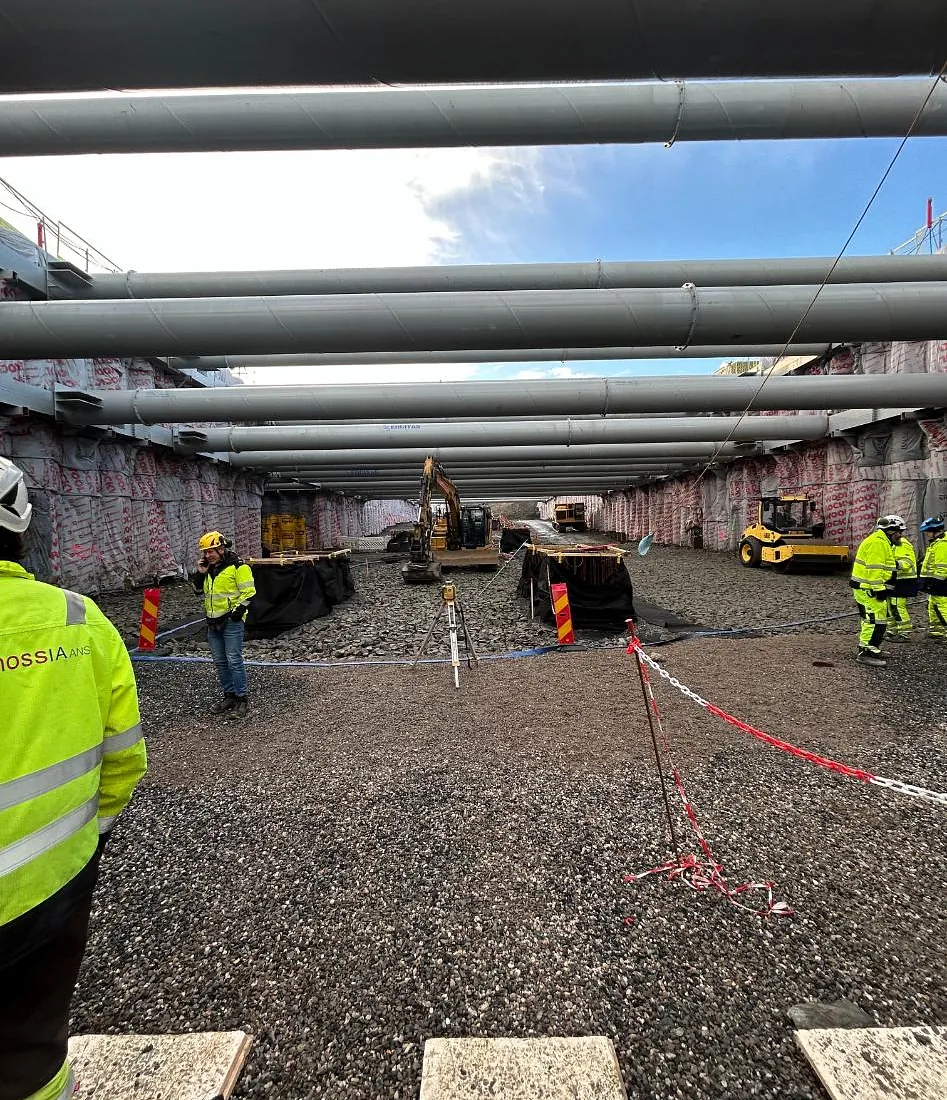 Anleggsområde med gravemaskiner og store vannrør over, flere arbeidere i refleksklær.